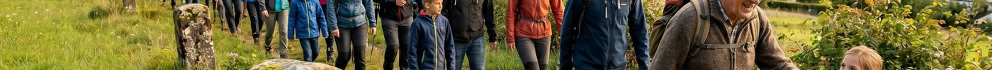 Eine Gruppe von Menschen unterschiedlichen Alters wandert bei einem traditionellen Grenzgang an einem alten Stein vorbei, im Hintergrund ein malerisches Dorf in hügeliger Landschaft.