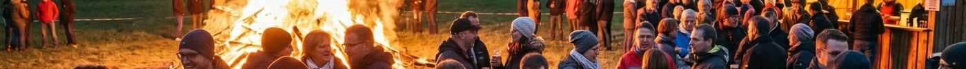 Eine große Gruppe Erwachsener steht am Abend gesellig um ein loderndes Maifeuer auf einer Wiese am Dorfrand; im Hintergrund sind beleuchtete Fachwerkhäuser und eine Kirche unter einem dämmerigen Abendhimmel zu sehen.