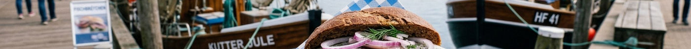 Eine Hand hält ein frisches Fischbrötchen mit Matjes und roten Zwiebeln vor der Kulisse eines malerischen Hafens mit Fischkuttern und einem Leuchtturm.