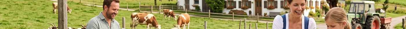 Eine Familie genießt einen sonnigen Tag auf einem idyllischen Bauernhof im Schwarzwald, umgeben von grünen Wiesen, Bergen, Kühen und Ziegen. Der Vater liest eine Karte, während die Mutter und Tochter am Tisch Teig kneten.
