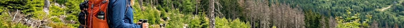 Wanderin mit orangefarbenem Rucksack blickt auf den Brocken im Harz mit der Brockenbahn und regenerierendem Wald im Hintergrund.