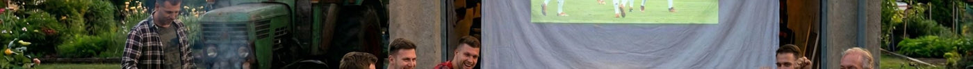 Eine Gruppe von Freunden schaut ein Fußball-Länderspiel auf einer improvisierten Leinwand vor einer Garage im Dorf; im Vordergrund sitzt ein Fan im weißen Podolski-Trikot mit der Nummer 10, daneben wird gegrillt.