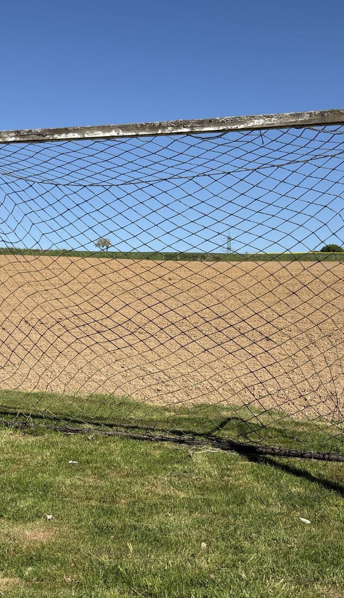 Ein altes, verwittertes Fußballtor mit Netz steht auf einem grünen Rasenstreifen vor einem frisch gepflügten, braunen Acker unter strahlend blauem Himmel.