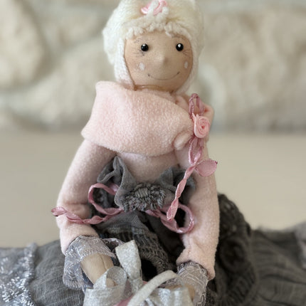 Zauberhafte, handgemachte Stoffpuppe mit rosa Mütze und Schal, einem grauen Spitzenrock und einem rosa Geschenk in den Händen, sitzend auf einem weißen Regal vor einer hellen Natursteinwand.