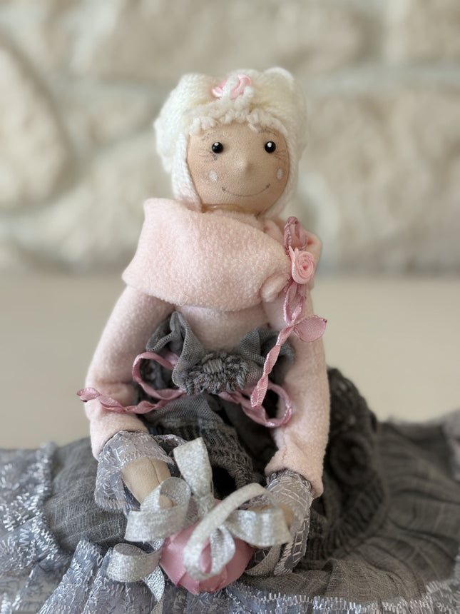 Zauberhafte, handgemachte Stoffpuppe mit rosa Mütze und Schal, einem grauen Spitzenrock und einem rosa Geschenk in den Händen, sitzend auf einem weißen Regal vor einer hellen Natursteinwand.