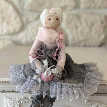 Zauberhafte, handgemachte Stoffpuppe mit rosa Mütze und Schal, einem grauen Spitzenrock und einem rosa Geschenk in den Händen, sitzend auf einem weißen Regal vor einer hellen Natursteinwand.