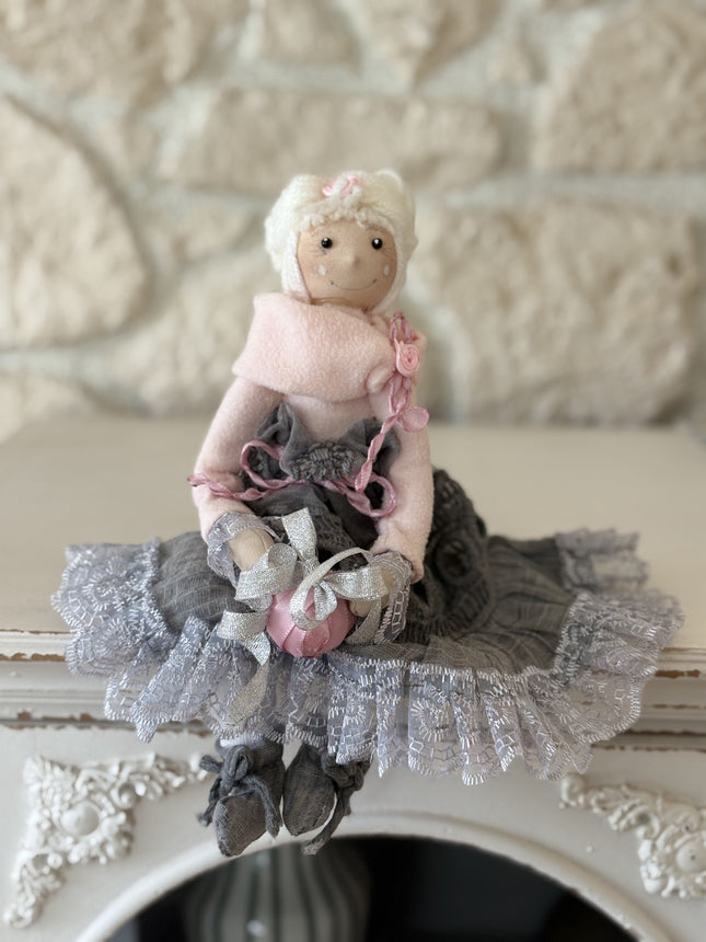 Zauberhafte, handgemachte Stoffpuppe mit rosa Mütze und Schal, einem grauen Spitzenrock und einem rosa Geschenk in den Händen, sitzend auf einem weißen Regal vor einer hellen Natursteinwand.