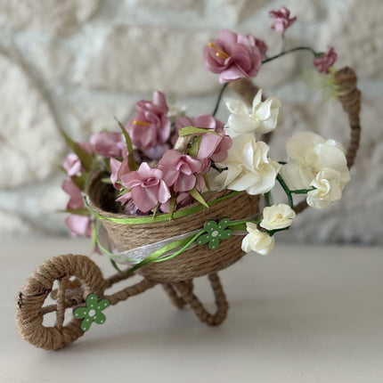 Handgefertigte, dekorative Miniatur-Schubkarre aus natürlicher Kordel, gefüllt mit einem Arrangement aus rosa, weißen und cremefarbenen Kunstblüten und Satinbändern. Das Rad ist mit einer kleinen, grünen Holzblume verziert.