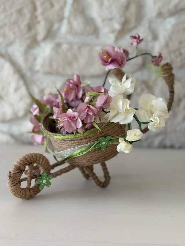 Handgefertigte, dekorative Miniatur-Schubkarre aus natürlicher Kordel, gefüllt mit einem Arrangement aus rosa, weißen und cremefarbenen Kunstblüten und Satinbändern. Das Rad ist mit einer kleinen, grünen Holzblume verziert.