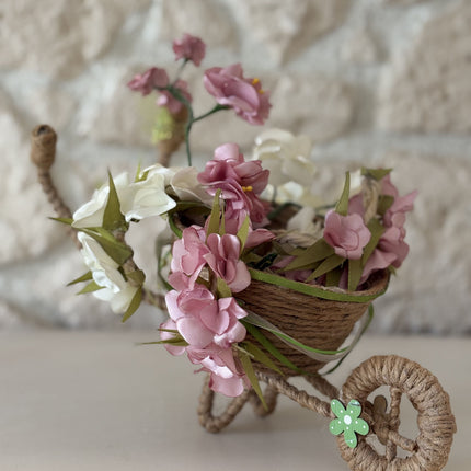 Handgefertigte, dekorative Miniatur-Schubkarre aus natürlicher Kordel, gefüllt mit einem Arrangement aus rosa, weißen und cremefarbenen Kunstblüten und Satinbändern. Das Rad ist mit einer kleinen, grünen Holzblume verziert.