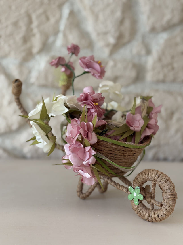 Handgefertigte, dekorative Miniatur-Schubkarre aus natürlicher Kordel, gefüllt mit einem Arrangement aus rosa, weißen und cremefarbenen Kunstblüten und Satinbändern. Das Rad ist mit einer kleinen, grünen Holzblume verziert.