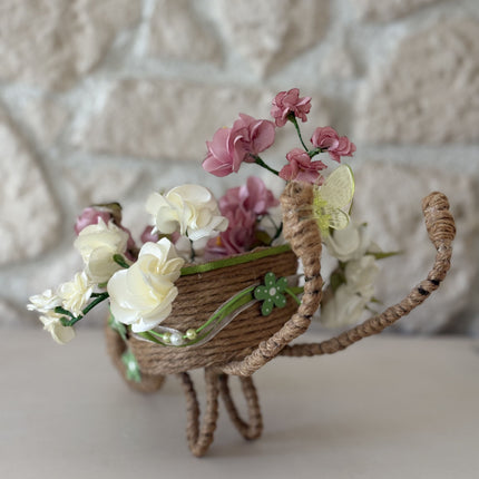 Handgefertigte, dekorative Miniatur-Schubkarre aus natürlicher Kordel, gefüllt mit einem Arrangement aus rosa, weißen und cremefarbenen Kunstblüten und Satinbändern. Das Rad ist mit einer kleinen, grünen Holzblume verziert.