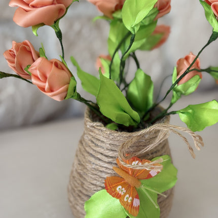 Nahaufnahme der handgefertigten Kunstrosen in Apricot. Die filigranen Blütenköpfe und das frische grüne Blattwerk sind deutlich zu erkennen. Der rustikale Jutetopf, umwickelt mit Schnur, ist im unteren Bereich sichtbar. Der Hintergrund ist eine hellbraune Natursteinwand, leicht unscharf.