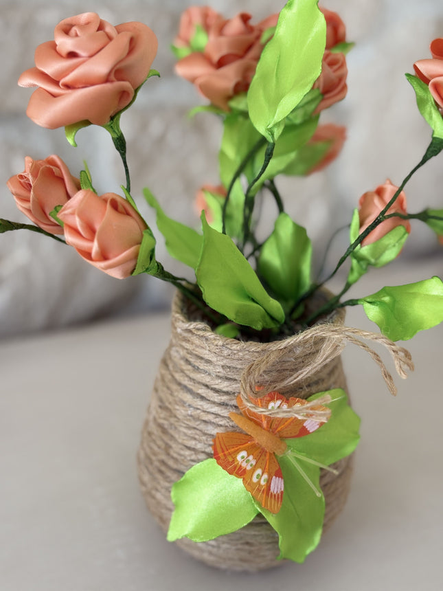 Nahaufnahme der handgefertigten Kunstrosen in Apricot. Die filigranen Blütenköpfe und das frische grüne Blattwerk sind deutlich zu erkennen. Der rustikale Jutetopf, umwickelt mit Schnur, ist im unteren Bereich sichtbar. Der Hintergrund ist eine hellbraune Natursteinwand, leicht unscharf.