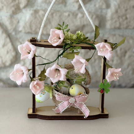 Handgemachte Osterdekoration in einem hellen Holzrahmen. Im Zentrum steht ein geflochtenes Körbchen mit bunt bemalten Ostereiern, verziert mit einer rosa Schleife. Umrahmt wird das Körbchen von zarten rosa Stoffblüten und grünen Blättern. Eine weiße Kordel dient als Aufhängung.