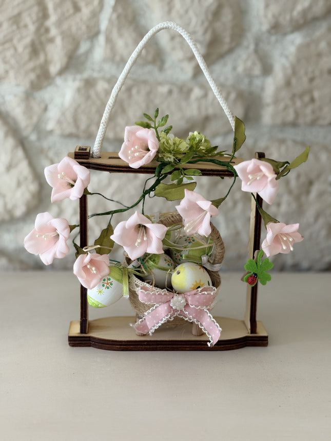Handgemachte Osterdekoration in einem hellen Holzrahmen. Im Zentrum steht ein geflochtenes Körbchen mit bunt bemalten Ostereiern, verziert mit einer rosa Schleife. Umrahmt wird das Körbchen von zarten rosa Stoffblüten und grünen Blättern. Eine weiße Kordel dient als Aufhängung.