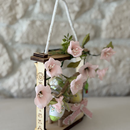 Handgemachte Osterdekoration in einem hellen Holzrahmen. Im Zentrum steht ein geflochtenes Körbchen mit bunt bemalten Ostereiern, verziert mit einer rosa Schleife. Umrahmt wird das Körbchen von zarten rosa Stoffblüten und grünen Blättern. Eine weiße Kordel dient als Aufhängung.