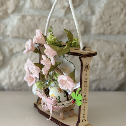 Handgemachte Osterdekoration in einem hellen Holzrahmen. Im Zentrum steht ein geflochtenes Körbchen mit bunt bemalten Ostereiern, verziert mit einer rosa Schleife. Umrahmt wird das Körbchen von zarten rosa Stoffblüten und grünen Blättern. Eine weiße Kordel dient als Aufhängung.