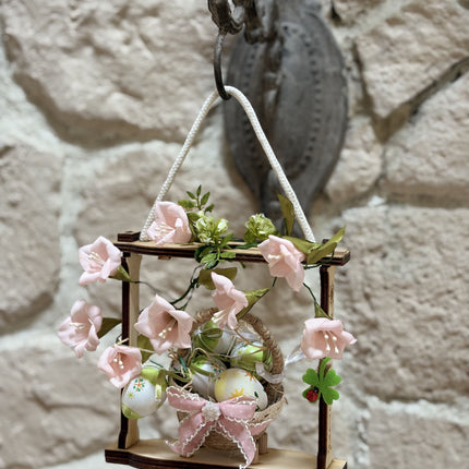 Handgemachte Osterdekoration in einem hellen Holzrahmen. Im Zentrum steht ein geflochtenes Körbchen mit bunt bemalten Ostereiern, verziert mit einer rosa Schleife. Umrahmt wird das Körbchen von zarten rosa Stoffblüten und grünen Blättern. Eine weiße Kordel dient als Aufhängung.