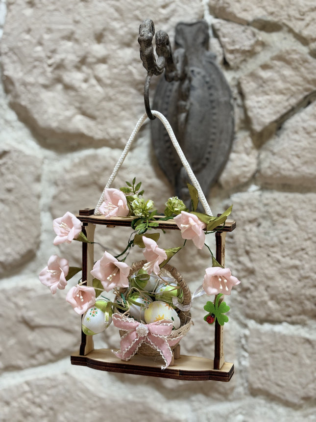 Handgemachte Osterdekoration in einem hellen Holzrahmen. Im Zentrum steht ein geflochtenes Körbchen mit bunt bemalten Ostereiern, verziert mit einer rosa Schleife. Umrahmt wird das Körbchen von zarten rosa Stoffblüten und grünen Blättern. Eine weiße Kordel dient als Aufhängung.