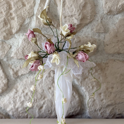 Handgefertigter, kleiner Deko-Schirm aus weißem Stoff, verziert mit künstlichen Rosen in Altrosa und Creme, feiner weißer Spitze und herabhängenden Blütenranken. Der Schirm hängt an einem rustikalen Metallhaken vor einer hellen Natursteinwand.
