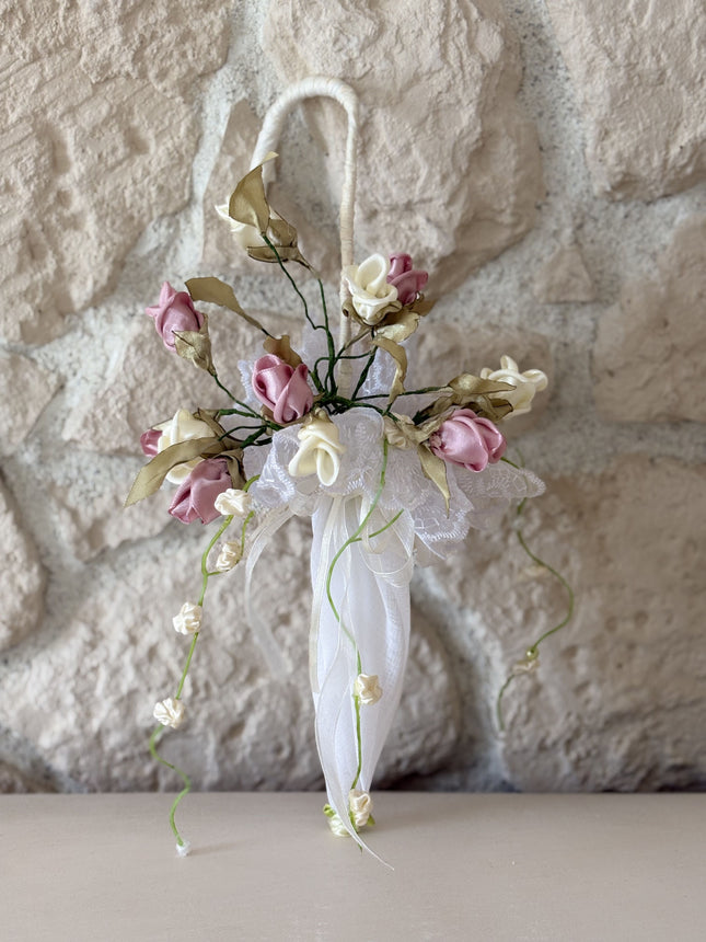 Handgefertigter, kleiner Deko-Schirm aus weißem Stoff, verziert mit künstlichen Rosen in Altrosa und Creme, feiner weißer Spitze und herabhängenden Blütenranken. Der Schirm hängt an einem rustikalen Metallhaken vor einer hellen Natursteinwand.