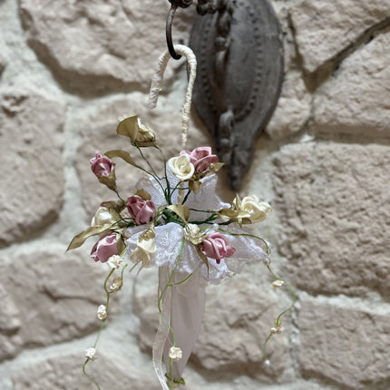Handgefertigter, kleiner Deko-Schirm aus weißem Stoff, verziert mit künstlichen Rosen in Altrosa und Creme, feiner weißer Spitze und herabhängenden Blütenranken. Der Schirm hängt an einem rustikalen Metallhaken vor einer hellen Natursteinwand.