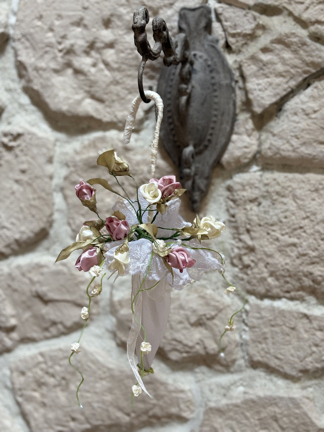 Handgefertigter, kleiner Deko-Schirm aus weißem Stoff, verziert mit künstlichen Rosen in Altrosa und Creme, feiner weißer Spitze und herabhängenden Blütenranken. Der Schirm hängt an einem rustikalen Metallhaken vor einer hellen Natursteinwand.