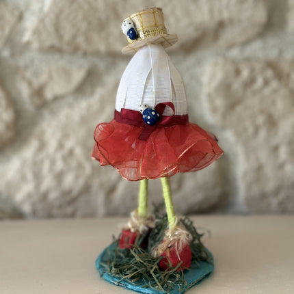 Ein aufrecht stehendes, handgefertigtes Deko-Kunstobjekt, das an eine menschliche Figur erinnert. Es besteht aus einem gewickelten weißen Oberkörper, einem hellgoldenen Zylinderhut mit Marienkäfer-Applikationen und einem roten Tüllrock. Die langen, drahtigen grünen Beine enden in roten Schuhen auf einer blauen Basis mit Strohdeko. Das Objekt ist detailliert und unique.