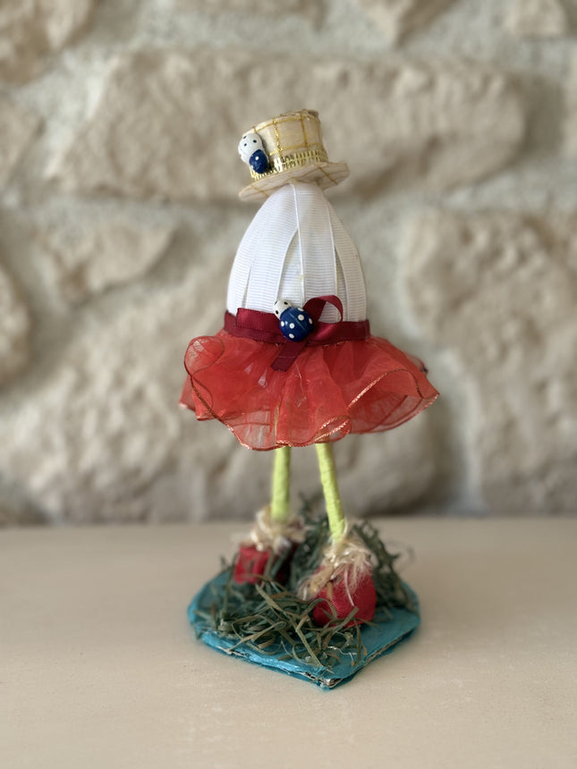 Ein aufrecht stehendes, handgefertigtes Deko-Kunstobjekt, das an eine menschliche Figur erinnert. Es besteht aus einem gewickelten weißen Oberkörper, einem hellgoldenen Zylinderhut mit Marienkäfer-Applikationen und einem roten Tüllrock. Die langen, drahtigen grünen Beine enden in roten Schuhen auf einer blauen Basis mit Strohdeko. Das Objekt ist detailliert und unique.