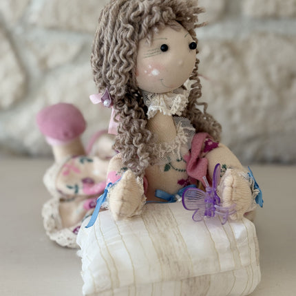 Foto einer handgemachten Stoffpuppe im Tilda-Stil mit braunem Lockenhaar und Blumenkleid, die auf einem kleinen, gestreiften Kissen liegt und eine lila Schmetterlingsklammer hält, vor dem Hintergrund einer rustikalen Steinwand.