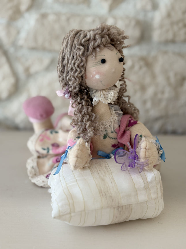 Foto einer handgemachten Stoffpuppe im Tilda-Stil mit braunem Lockenhaar und Blumenkleid, die auf einem kleinen, gestreiften Kissen liegt und eine lila Schmetterlingsklammer hält, vor dem Hintergrund einer rustikalen Steinwand.
