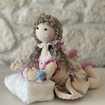 Foto einer handgemachten Stoffpuppe im Tilda-Stil mit braunem Lockenhaar und Blumenkleid, die auf einem kleinen, gestreiften Kissen liegt und eine lila Schmetterlingsklammer hält, vor dem Hintergrund einer rustikalen Steinwand.