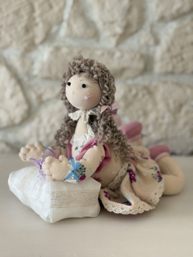 Foto einer handgemachten Stoffpuppe im Tilda-Stil mit braunem Lockenhaar und Blumenkleid, die auf einem kleinen, gestreiften Kissen liegt und eine lila Schmetterlingsklammer hält, vor dem Hintergrund einer rustikalen Steinwand.